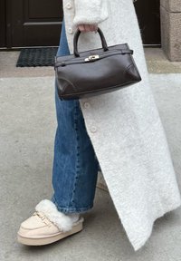 Personne portant un jean bleu, des chaussures à enfiler beiges doublées en peau lainée, et un long cardigan blanc tenant un sac à main noir près d'une porte.