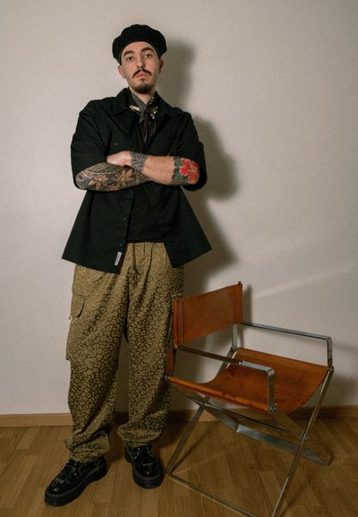 Camisa negra de manga corta, camiseta negra, pantalones cargo de estampado de leopardo marrón, zapatos negros, tatuajes visibles en los brazos, suelo de madera, silla de metal.