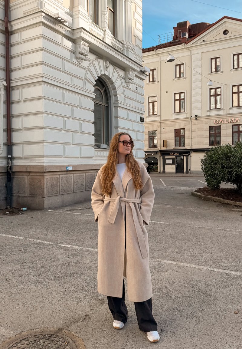 Femme aux longs cheveux et lunettes de soleil, portant un manteau beige ceinturé, un pantalon noir et des chaussures blanches, debout dans une rue de la ville près de bâtiments classiques.