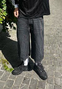 Fekete farmer culotte sort, elhalványodott textúrával, fekete tornacipővel és fehér zoknival párosítva. A háttérben kőburkolat és zöld növényzet látható.