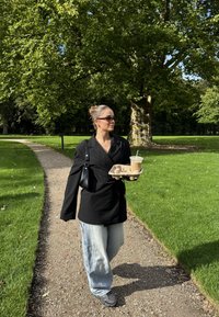 Svart oversized blazer, lyseblå baggy jeans, svarte joggesko. Holder en kaffekopp i en pappbrett langs en buet grussti i en park.