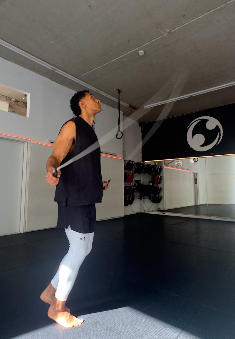 Hombre con camiseta negra sin mangas y mallas blancas saltando la cuerda descalzo en un gimnasio con pared de espejos y equipo de artes marciales en estantes.