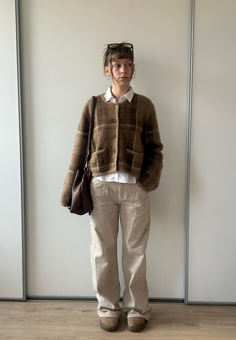 Jeune femme debout à l'intérieur portant un cardigan à carreaux marron, chemise blanche, pantalon beige, bottes marron, portant un sac à bandoulière marron, avec des lunettes sur la tête.