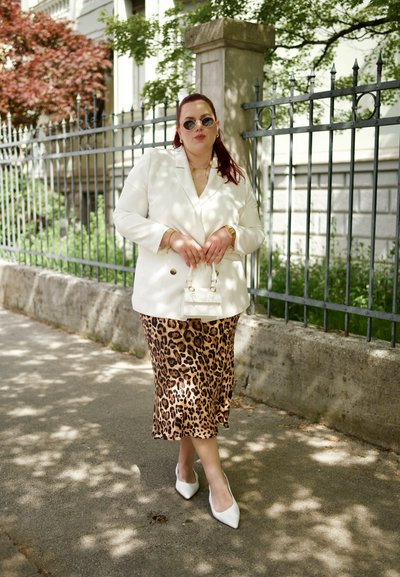 Blazer blanco, falda midi de estampado de leopardo, tacones blancos de punta. Bolso de mano blanco pequeño. Gafas de sol y joyería dorada complementan el atuendo.