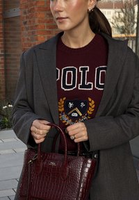 Bolsa de couro com textura de crocodilo na cor bordeaux, com alças duplas, ferragens douradas e logotipo em relevo. Combinada com um blazer cinza e um suéter em bordô.