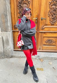 Maglione grigio oversize, sciarpa frangiata rossa, gonna nera lucida, collant rossi, stivali neri chunky e una borsa con motivo fragola. Sfondo con porta in legno.