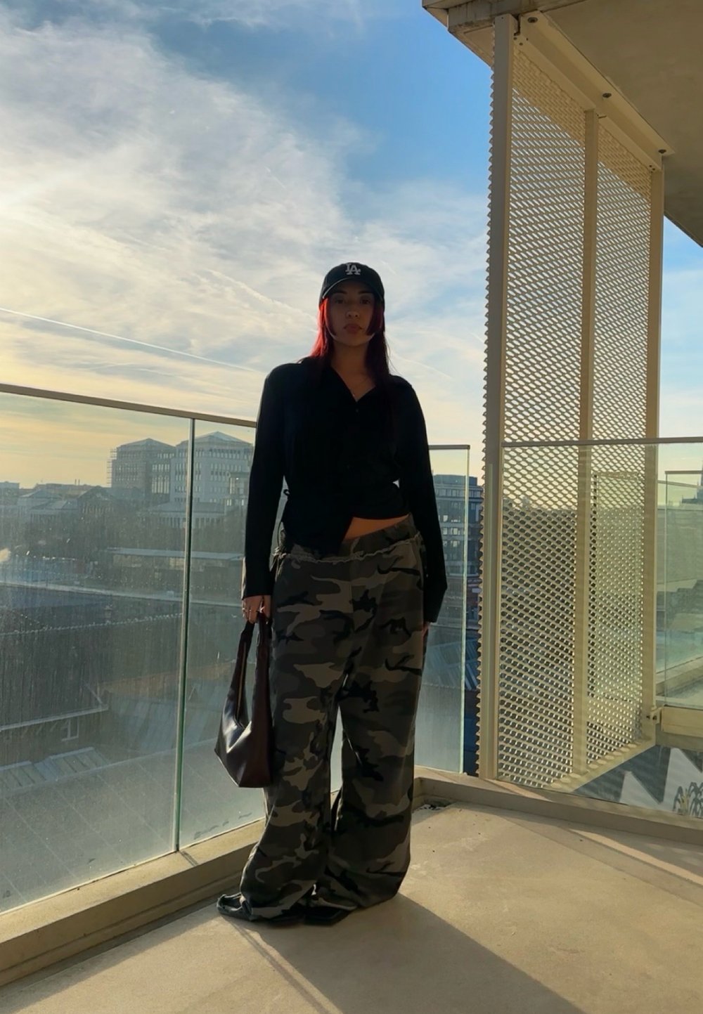 Black button-up shirt, camo wide-leg trousers, black cap, and brown tote bag. Standing on a balcony with glass railing under a blue sky.