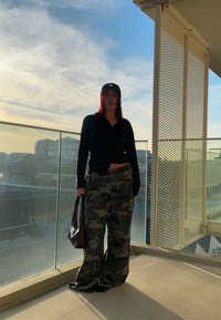 Black button-up shirt, camo wide-leg trousers, black cap, and brown tote bag. Standing on a balcony with glass railing under a blue sky.