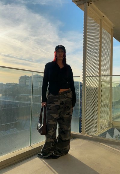 Camisa negra de botones, pantalones anchos de camuflaje, gorra negra y bolsa de mano marrón. De pie en un balcón con barandilla de vidrio bajo un cielo azul.