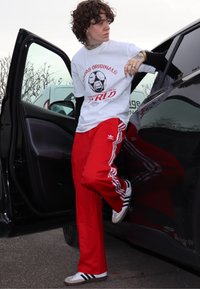 Hvit t-skjorte med grafisk trykk, røde Adidas treningsbukser med hvite striper, og hvite joggesko med svarte detaljer, stående ved siden av en bil.