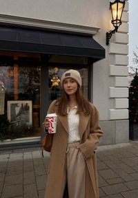 Femme en manteau beige et bonnet en laine tenant un gobelet de café à thème festif, debout sur le trottoir près d'un magasin décoré avec un arbre de Noël et une lanterne.