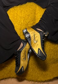Zapatillas Nike negras y doradas con un upper texturizado, diseño elegante y amortiguación visible. Colocadas sobre un fondo amarillo esponjoso.