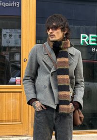 Manteau gris croisé sur un pull marron, accompagné d'une écharpe rayée noire, marron et verte. Jean foncé et sac marron.
