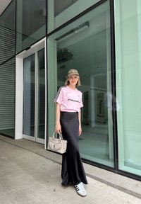 Pink adidas T-shirt med sorte striber, lang sort nederdel og grå sneakers. Holder en grå håndtaske. Står på en betonoverflade nær glasdøre.