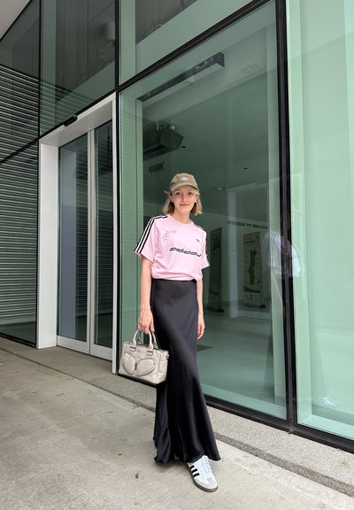 Camiseta rosa de adidas con rayas negras, falda larga negra y zapatillas grises. Sosteniendo un bolso gris. De pie sobre una superficie de hormigón cerca de puertas de cristal.