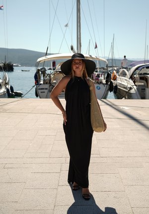 Vestido maxi negro con diseño de cuello halter, sombrero negro de ala ancha y un bolso de paja trenzada, de pie sobre un pavimento de piedra cerca de botes.