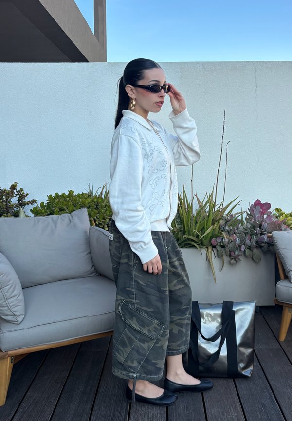 Kvinne som justerer svarte solbriller, iført hvit cardigan og kamuflasjete vide bukser, står ved siden av sofa og blank svart totebag utendørs.