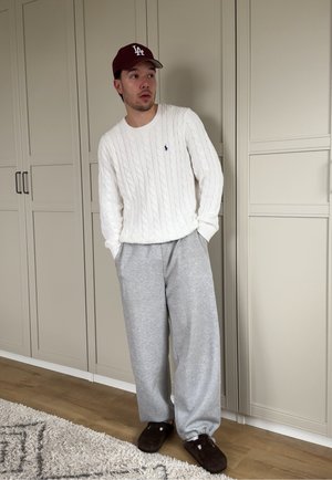 Joven con un suéter blanco de punto trenzado, pantalones deportivos grises, zapatillas marrones y una gorra de béisbol granate, de pie en el interior junto a armarios beige.