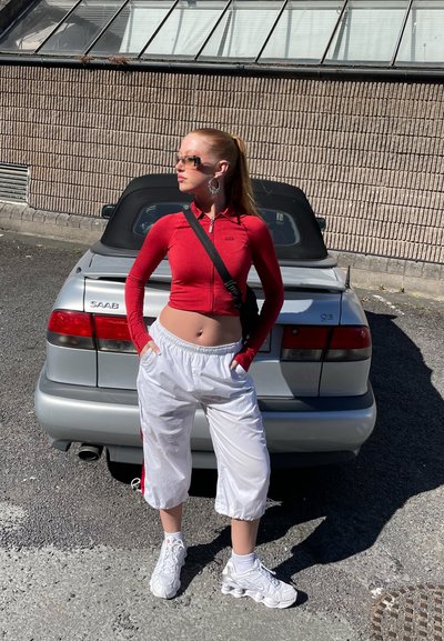 Sudadera roja de manga larga con cremallera, pantalones de chándal cortos blancos, zapatillas metálicas; de pie frente a un coche convertible plateado.