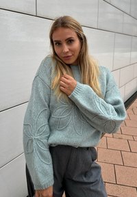 Vaaleansininen neulepusero, jossa on kukkakirjontaa hihoissa, rento istuvuus ja ribbireunat, yhdistettynä korkeavyötäröisiin harmaisiin housuihin.