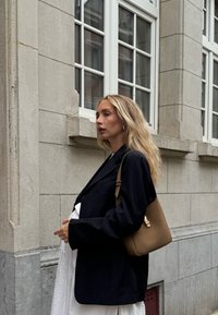 Blazer preto, top branco e bolsa bege. A bolsa tem um fecho dourado e o conjunto contrasta com um edifício de pedra clara.