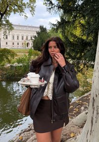 Barna bőrkabát cipzárral, fehér csipke felső és fekete miniszoknya. Egy tálcát tart, rajta két kávéscsésze, egy telefon és fülhallgatók.