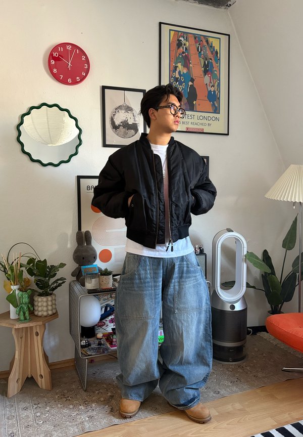 Ung mand iført oversized jeans og sort jakke stående i et dekoreret opholdsstue med planter, kunstværker, et rødt ur og en hvid luftrenser.