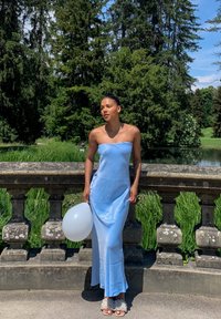 Robe en satin bleu clair, sans manches, avec un bustier ajusté et une jupe fluide. Le mannequin tient un ballon blanc et porte des chaussures ouvertes avec des accents en perles.