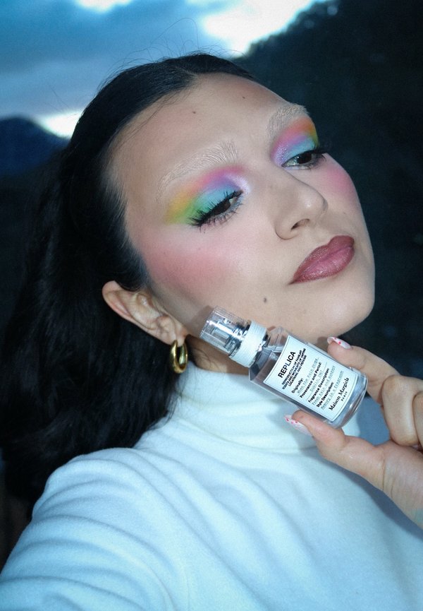 Woman with rainbow eyeshadow, gold hoop earrings, and white turtleneck holding Maison Margiela Replica perfume near her cheek.