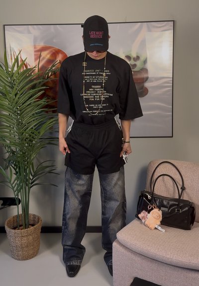 Camiseta negra de gran tamaño con texto gráfico, pantalones cortos negros, jeans de mezclilla desgastados, gorra negra y bolso negro con llaveros de peluche.