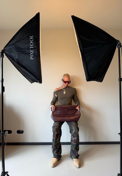 Bolso burdeos con textura de cocodrilo, con solapa y hebilla, sostenido por una persona que lleva una blusa ceñida color oliva y pantalones de camuflaje, frente a una pared neutral.