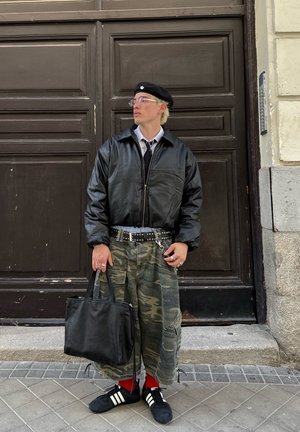 Juodas odinis bomber tipo švarkas, balta marškinė su apykakle, kamufliažo kroviniai šortai, juodas diržas su siaurais metaliniais kaiščiais, juodas rankinis krepšys, raudonos kojinės ir juodi sportbačiai.