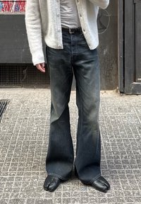 Φλάρε τζιν σε σκούρο μπλε τζιν, φορεμένα με ανοιχτό γκρι πουκάμισο και λευκό πλεκτό ανοιχτό. Μαύρα παπούτσια με διαχωρισμό στα δάχτυλα ολοκληρώνουν το σύνολο.