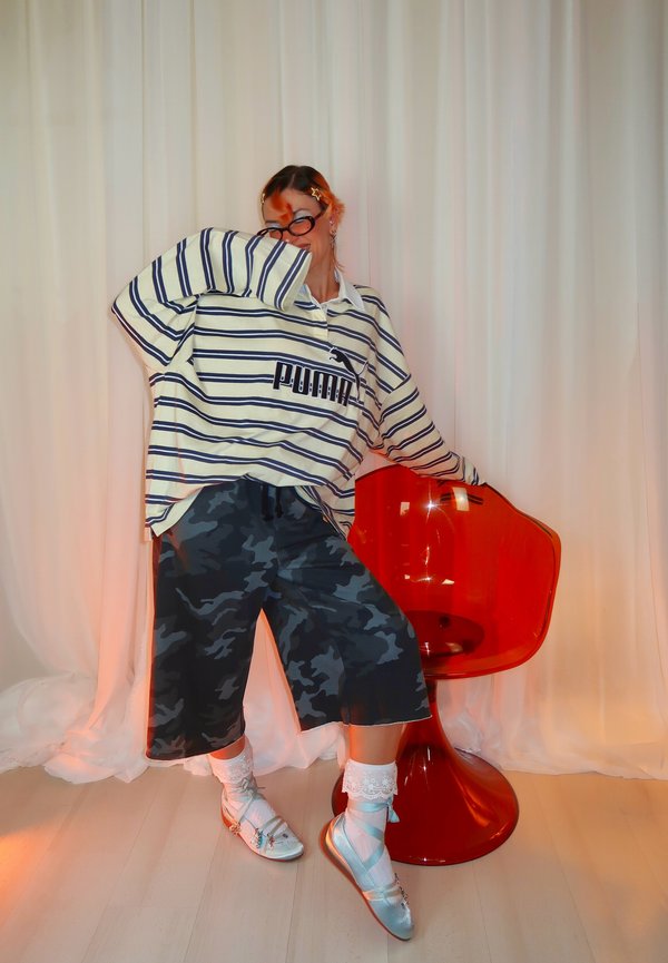 Personne portant une chemise rayée Puma oversize, un short camouflage, des chaussettes blanches et des chaussures argentées, posant à côté d'une chaise rouge translucide devant des rideaux blancs.