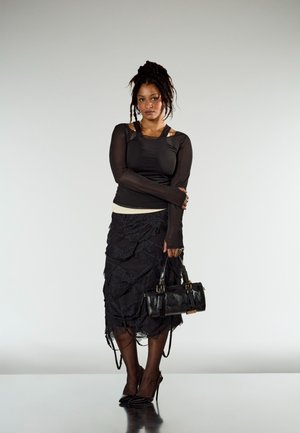 A black sheer long-sleeve top layered over a cream tank top, paired with a black ruffled skirt. Accessorised with a black handbag and pointed heels.