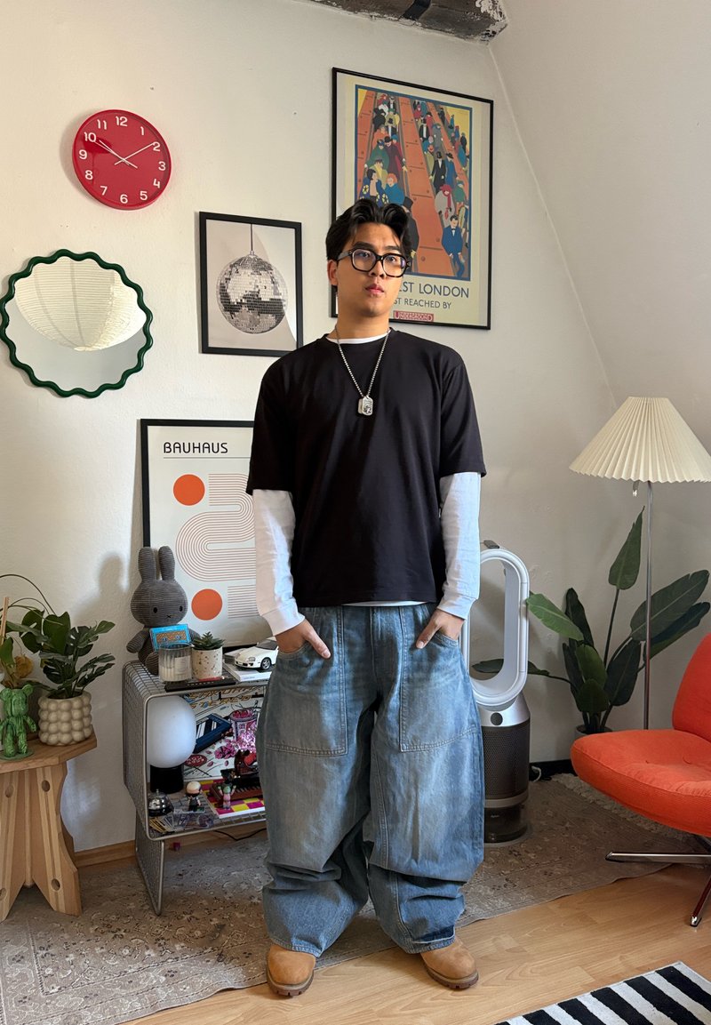 Jeune personne portant des lunettes, habillée d'une superposition de chemises noires et blanches et d'un jean ample, se tient dans une pièce décorée avec des plantes, des affiches et une chaise orange.