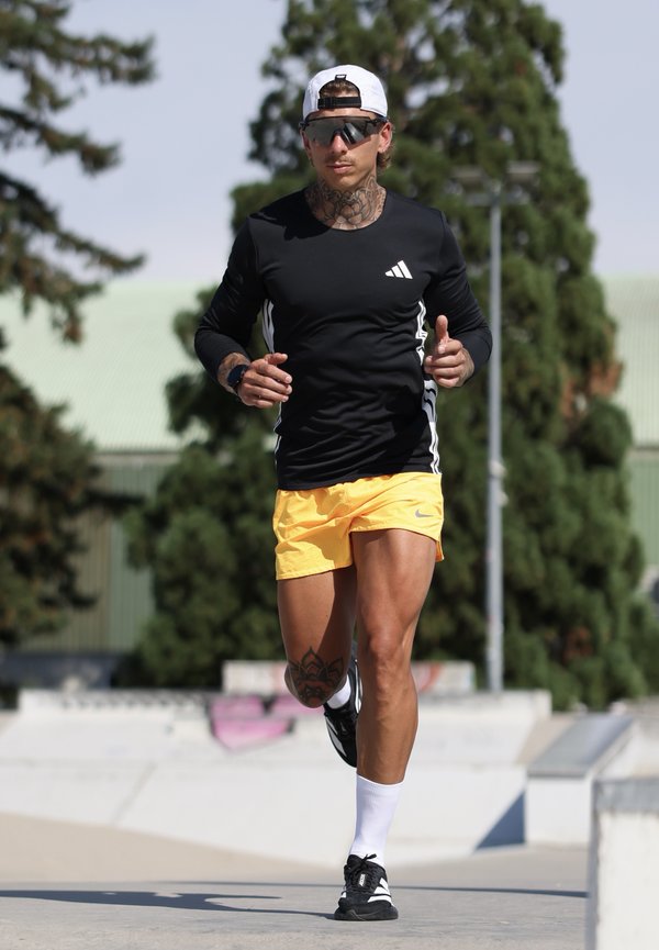 Homme tatoué portant des lunettes de soleil, un t-shirt à manches longues noir, un short jaune et une casquette blanche, courant en plein air sur un chemin pavé.