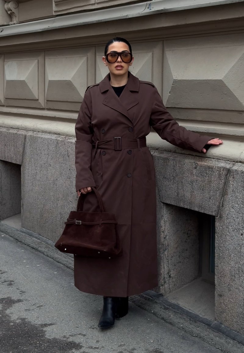 Femme portant un long trench-coat marron, de grandes lunettes de soleil, des bottes noires, tenant un sac à main marron, debout contre un mur en pierre texturé dans une rue de la ville.
