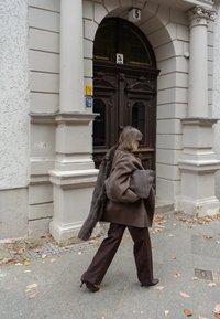 Barna gyapjúkabát szőrmés részlettel, sötétbarna nadrág, szürke táskát visel, világos színű épület mellett halad el, amelynek faajtaja van.