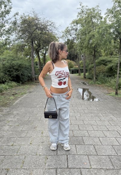 Camiseta sin mangas blanca con estampado de "Cherry Bomb" y gráfico de cereza, pantalones de chándal grises, bolso mini negro y zapatillas grises, al aire libre en un camino pavimentado.