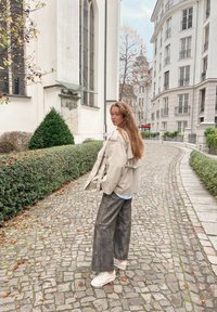 Veste beige oversize avec détails à volants, superposée sur une chemise bleu clair, associée à un pantalon large foncé et des baskets blanches. Chemin pavé.