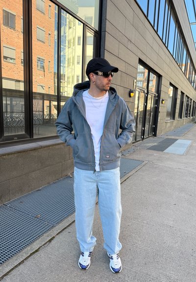 Sudadera gris con cremallera sobre una camiseta blanca, jeans de mezclilla azul claro y zapatos deportivos blancos con acentos en azul marino. Una gorra negra y gafas de sol completan el look.