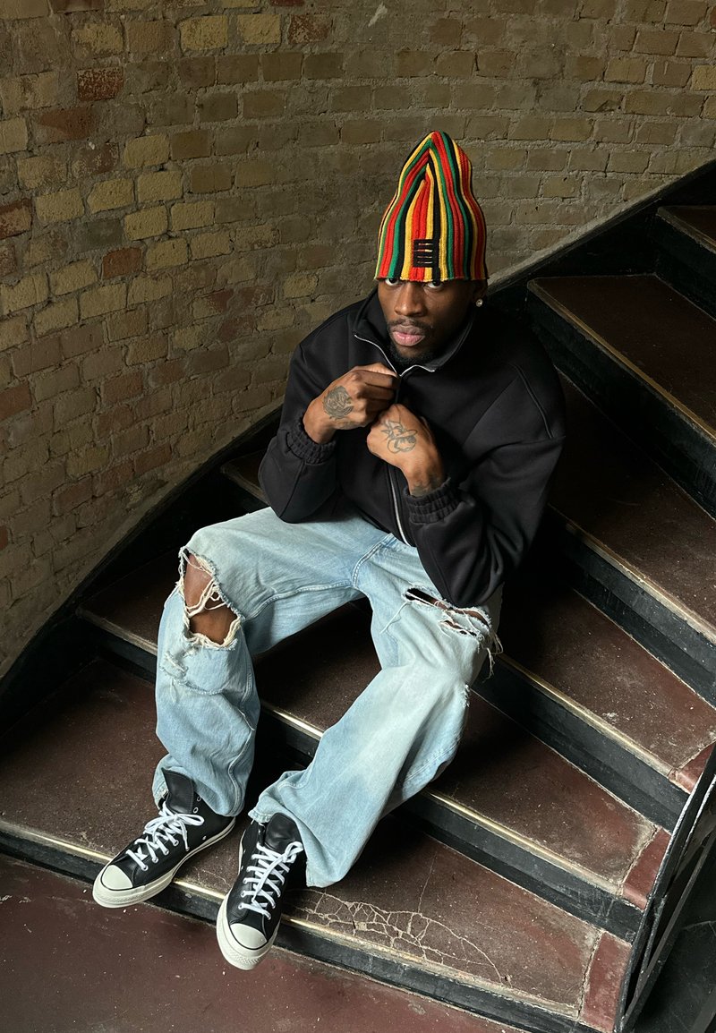 Homme assis sur des escaliers portant une veste noire, un jean bleu clair déchiré, des baskets noires et un grand bonnet en tricot rayé multicolore.
