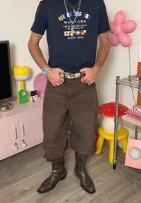 Mand iført en mørkeblå grafisk T-shirt, brune bukser stoppet ned i cowboystøvler, står i et farverigt rum med legende dekorationer og hylder.