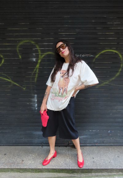 Mujer con gafas de sol, camiseta de ciervo oversize, pantalones cortos negros, zapatos rojos y un clutch rojo, de pie frente a una persiana con grafitis.