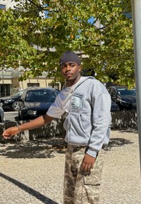 Hanorac gri cu fermoar, cu un buzunar frontal, purtat peste un tricou alb, asortat cu pantaloni cargo bej cu model de camuflaj. Înconjurat de frunziș verde.