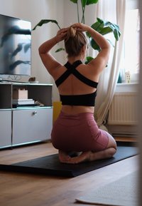 Top de desporto preto com design cruzado nas costas, calções rosa de cintura alta, ajoelhando-se sobre um tapete de yoga preto numa sala iluminada com plantas e uma televisão.