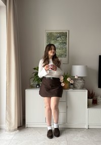 Brun miniskjørt med nålestriper, hvit langermet topp, mørke loafers og hvite sokker med bølgete kanter; innendørs setting med planter og dekor.