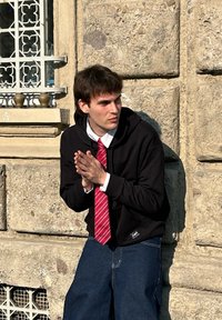 Fekete kapucnis pulóvert és piros csíkozott nyakkendőt viselő személy egy texturált kőfal előtt áll, összefont kezekkel, laza kék farmerban.