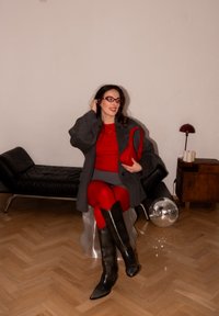 Femme en robe rouge, manteau gris, bottes noires et lunettes rouges assise sur un tabouret argenté, tenant un sac rouge dans une pièce moderne.